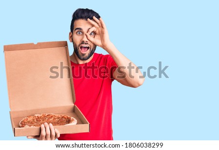 Young handsome man with beard holding delivery cardboard with italian pizza smiling happy doing ok sign with hand on eye looking through fingers 
