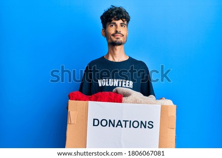 Young hispanic man holding donation box with clothes smiling looking to the side and staring away thinking. 