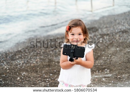 Caucasian small girl with selfie stick on the seaside. Taking photo, recording vlog, video call