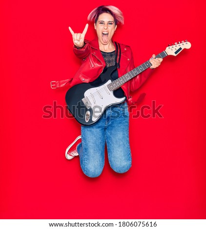 Young beautiful guitarist woman with pink short hair playing electric guitar smiling happy. Jumping with smile on face doing rocker sign with fingers over isolated red background