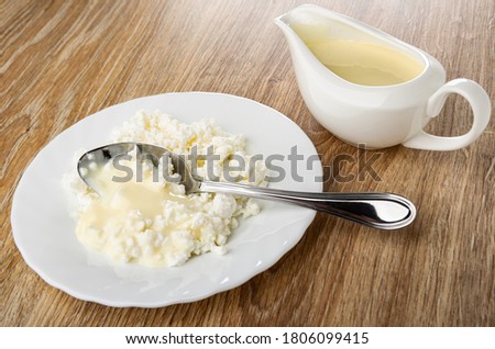 Spoon in white glass plate with grainy cottage cheese poured condensed milk, sauce boat with milk on wooden table Royalty-Free Stock Photo #1806099415