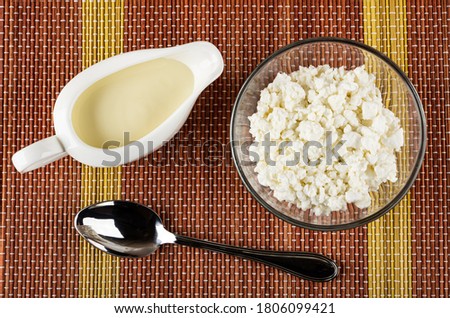 Sauce boat with condensed milk, transparent glass bowl with grainy cottage cheese, spoon on bamboo mat. Top view Royalty-Free Stock Photo #1806099421