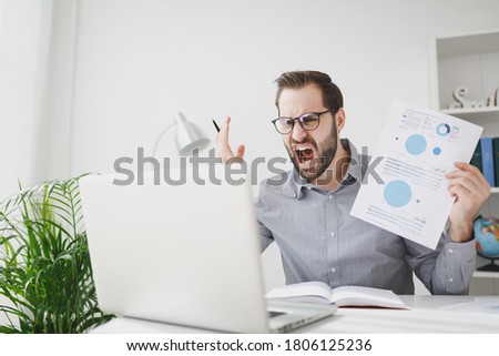 Angry business man in gray shirt glasses sit at desk in light office work on laptop make video call hold paper document scream swearing on white wall background. Achievement business career concept.
