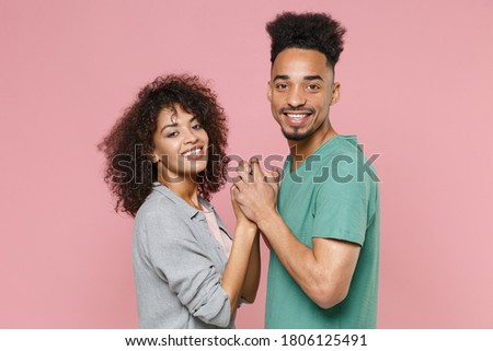 Side view of smiling young african american couple two friends woman man in gray green casual clothes posing holding hands looking camera isolated on pastel pink color wall background studio portrait