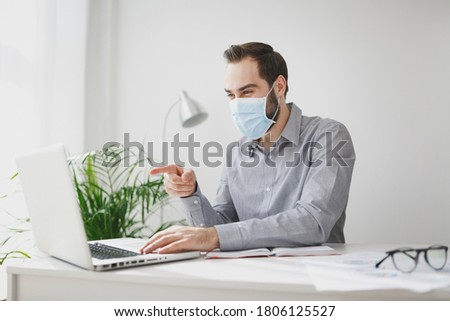 Young business man in gray shirt sterile face mask sit at desk in office work pointing finger on laptop pc computer making video call on white wall background. Achievement business career concept.