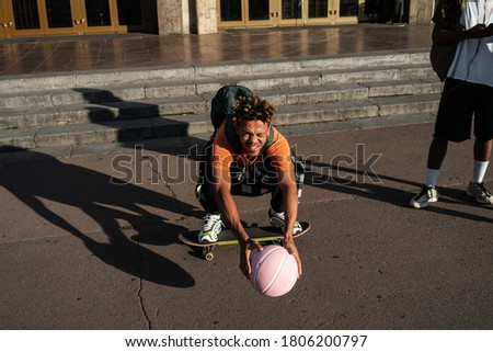 Photo of a young african optimistic teenage friends walking outdoors with ball and skateboard