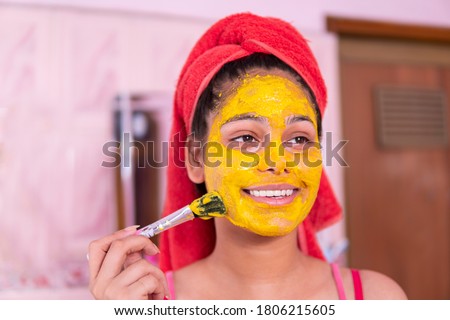Skin care - Beautiful girl applying Gram flour turmeric yellow face mask on face through brush. She is wearing red towel on head. Royalty-Free Stock Photo #1806215605
