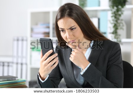 Suspicious executive woman checking smart phone sitting on a desk at the office Royalty-Free Stock Photo #1806233503