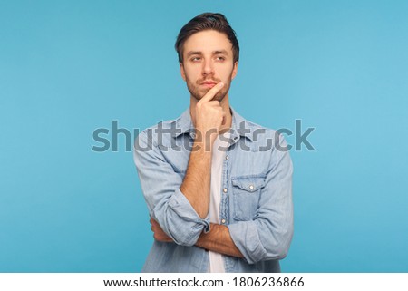 Hmm, let's think! Portrait of pensive handsome man in worker denim shirt touching chin while pondering plan, having doubts about difficult choice, not sure. studio shot isolated on blue background Royalty-Free Stock Photo #1806236866