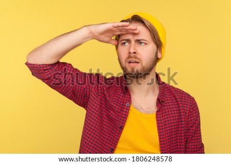 Far away vision. Hipster bearded guy in beanie hat and checkered shirt holding hand over eyes looking long distance with attentive view, future perspectives. studio shot isolated on yellow background