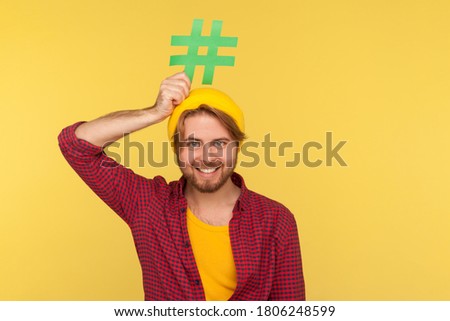 Hashtag, internet trends. Happy funny hipster guy in checkered shirt smiling and holding hash sign over head, symbol of viral message social network. indoor studio shot isolated on yellow background