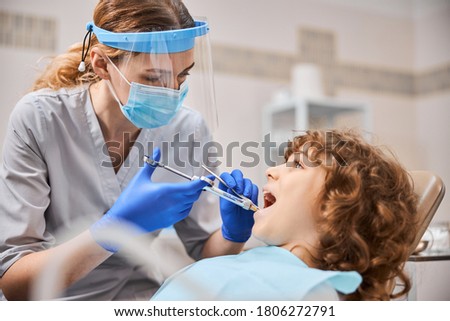 Focused fair-haired dentist wearing protective equipment and giving an anesthesia injection to kid