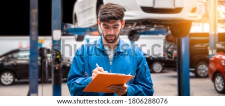 Mechanic in blue work wear uniform checks the vehicle maintenance checklist with blur lifted car in the background. Automobile repairing service, Professional occupation. Royalty-Free Stock Photo #1806288076