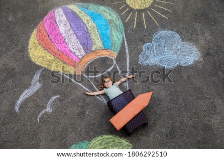 Happy little toddler girl flying in hot air balloon painted with colorful chalks in rainbow colors on ground or asphalt in summer. Cute child having fun. Creative leisure for kids.