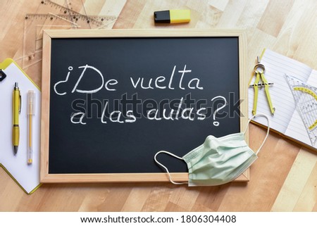 Blackboard with text "back to the classrooms?", school supplies and mask on a wooden table. Debate on face-to-face teaching after the coronavirus stop.