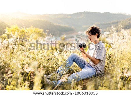 Happy young photographer sitting in the middle of the grass, taking pictures of the landscape, with a beautiful sunny day.
