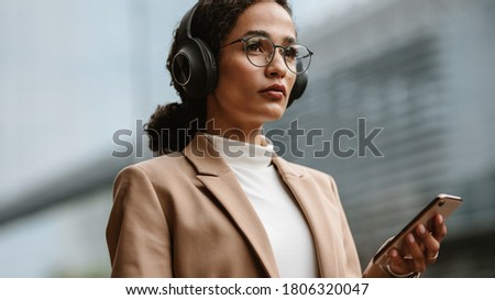Confident female executive listening to music on cell phone. Businesswoman wearing headphones and holding a phone while standing outdoors on city street. Royalty-Free Stock Photo #1806320047