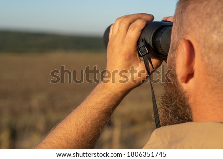 A bearded man looks through binoculars, close-up. The hunter uses binoculars to search