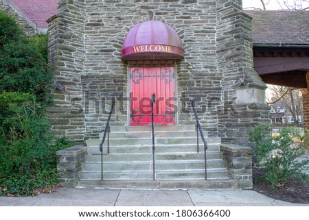 A Bright Red Door in a Cobblestone Wall With a Welcome Sign Above and Steps Leading Up to It and a Bush on the Side