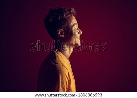 Profile view of happy young adult guy with smiling face wearing in casual stylish clothes, standing against dark red background with soft light, shadows and copy space