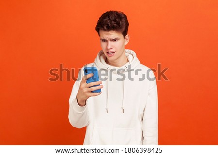 Wondered teenager in white casual sweatshirt with hood looking at smartphone screen with amazed face, bad connection. Indoor studio shot isolated on orange background