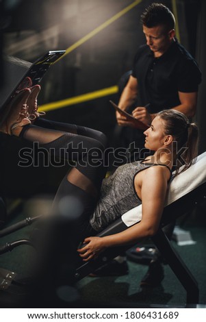 Shot of a muscular young woman in sportswear working out with personal trainer at the gym. She is doing exercises for her legs on leg press machine. Royalty-Free Stock Photo #1806431689