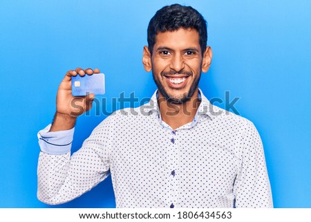 Young latin man holding credit card looking positive and happy standing and smiling with a confident smile showing teeth 