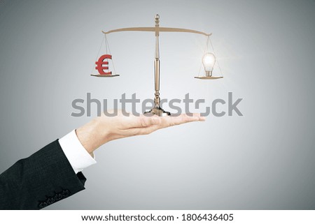 Hand holding golden scales with euro money symbol and lightbulb on gray background. Business and financial success concept.