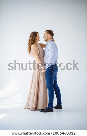 A beautiful curly pregnant girl in a long evening light  dress dancing with  her husband on a white background. She has a long evening dress and he is wearing festive blue shirt. Happy  Family Concept