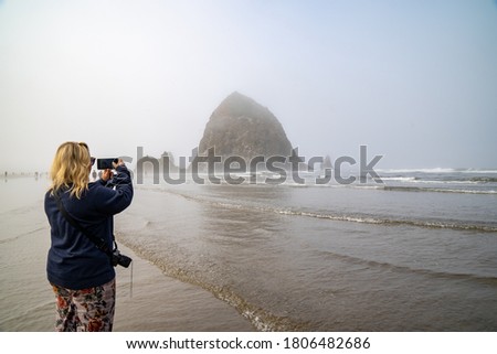 Blonde woman tourists takes photos with her phone of Haystack Rock in Cannon Beach Oregon