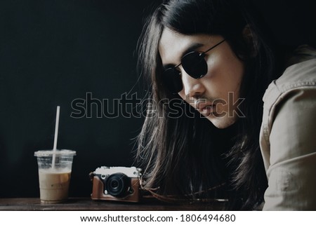 Closeup picture of white Asian men long hair, wear sunglasses  with his camera and a cup of coffee on black background. Light and shadow style. Photographer concept
