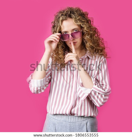 The young woman holds onto her glasses and makes a gesture with her hands - quietly. Secret, secret, isolated on pink background