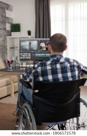 Videographer with handicap sitting on wheelchair doing color grading on film. Invalid film maker doing his business online, using high technology, sitting in his apartment, working remotely in special Royalty-Free Stock Photo #1806572578