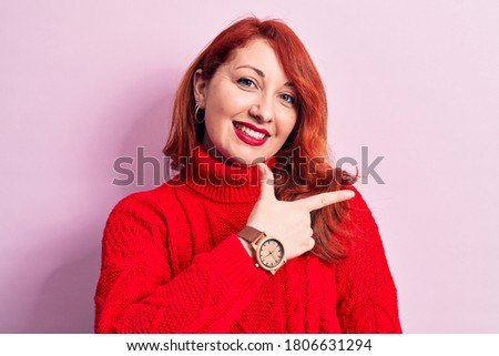 Young beautiful redhead woman wearing red casual turtleneck sweater over pink background smiling cheerful pointing with hand and finger up to the side