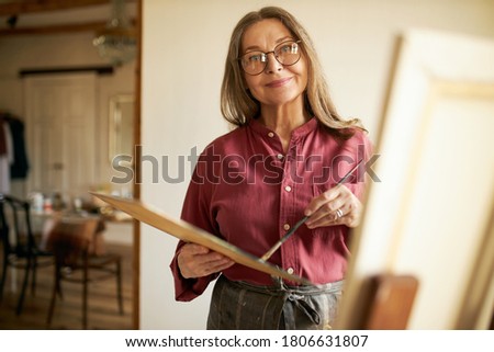 Long-haired middle aged female artist in spectacles standing in front of canvas holding brush and palette painting. Beautiful mature woman artist working in her studio. Creativity and inspiration