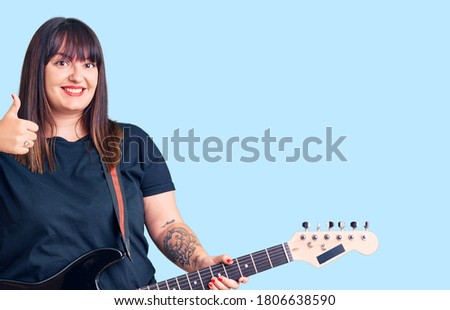 Young plus size woman playing electric guitar smiling happy and positive, thumb up doing excellent and approval sign 