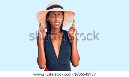 Young african american woman wearing swimsuit and summer hat gesturing finger crossed smiling with hope and eyes closed. luck and superstitious concept. 