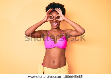 Young african american woman wearing bikini doing ok gesture like binoculars sticking tongue out, eyes looking through fingers. crazy expression. 