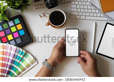 Top view of designer holding mock up smartphone in his workspace with Pantone swatches and office accessories 