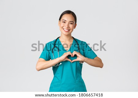 Covid-19, healthcare workers, pandemic concept. Lovely caring asian doctor, female nurse in scrubs showing heart gesture and smiling, taking care of patients with love, white background