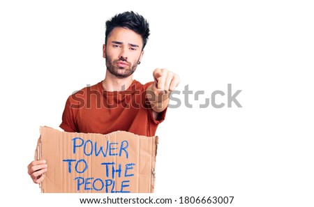 Young hispanic man holding power to the people banner pointing with finger to the camera and to you, confident gesture looking serious 