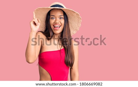 Young beautiful latin girl wearing swimwear and summer hat pointing finger up with successful idea. exited and happy. number one. 