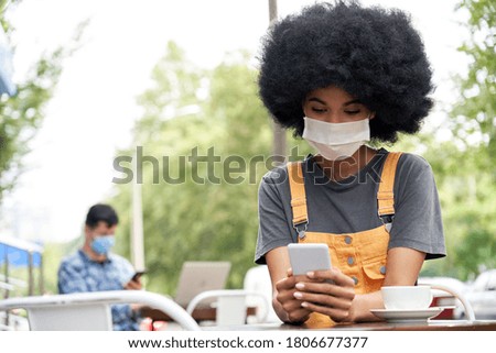 African American young hipster gen z female with Afro hair wearing face mask holding smart phone using mobile apps sitting at table in outdoor cafe. Social distancing everyday life and tech concept Royalty-Free Stock Photo #1806677377