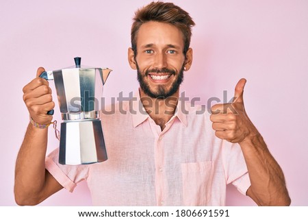 Handsome caucasian man with beard holding italian coffee maker smiling happy and positive, thumb up doing excellent and approval sign 