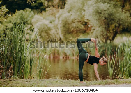 Happy skinny beautiful fit yoga woman in green leggings and black sport top is practicing yoga balance pose in outdoor by the lake. Fitness, sport, people and healthy lifestyle concept.