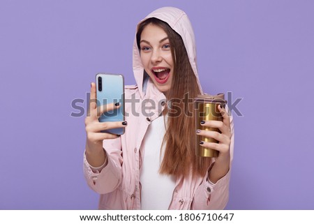 Attractive Caucasian girl making selfie against lilac background, being excited, keeping mouth widely opened, enjoying hot beverage, wearing pale pink raincoat.