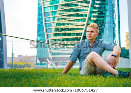  young man in a shirt rests on the grass in a business district with a view of glass skyscrapers