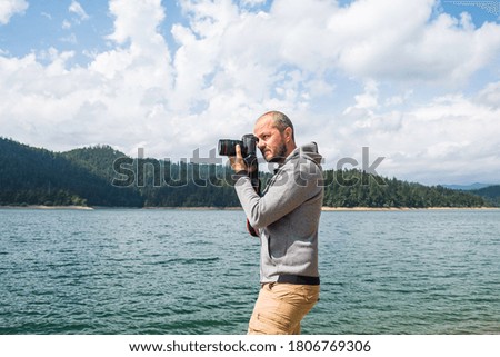 Photographer man in hoodie walking around lake with DSLR camera and shooting nature, half body portrait, landscape photography concept
