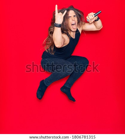 Young handsome heavy metal singer man with long hair and aggressive expression. Singing song using microphone jumping over isolated red background.