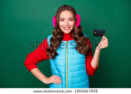 Photo of pretty lovely young lady smiling showing credit card hand hip bank promotion profitable deposit saving wear pink ear warmers blue vest red sweater isolated green color background